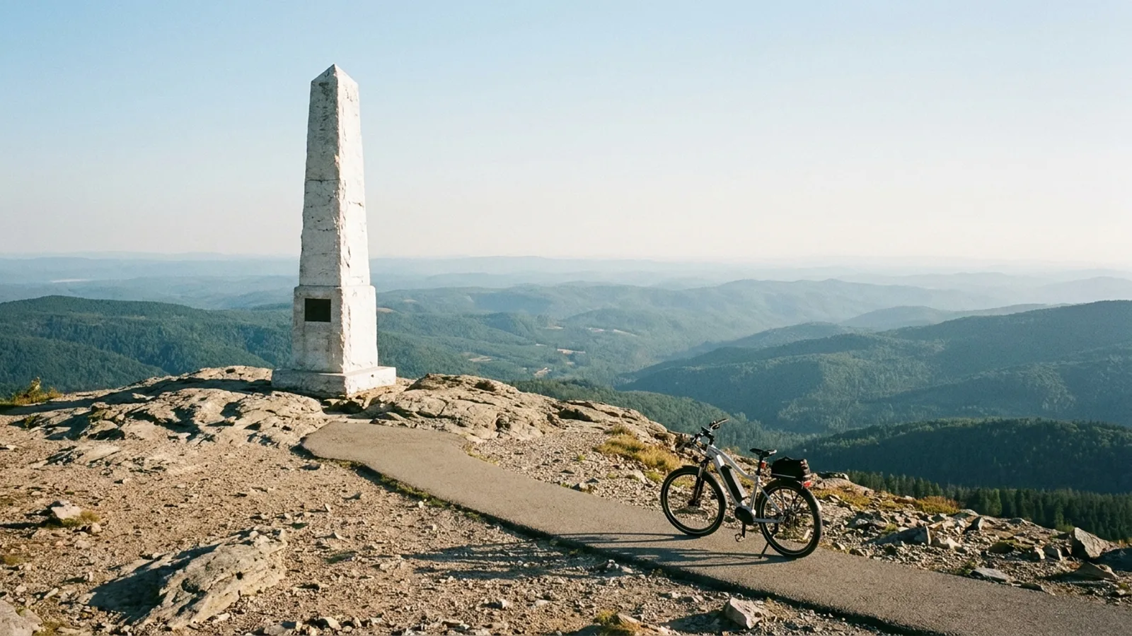 Electric bike on a paved path leading to a tall white stone obelisk monument on a bare rocky mountain summit with long views over forested ridges