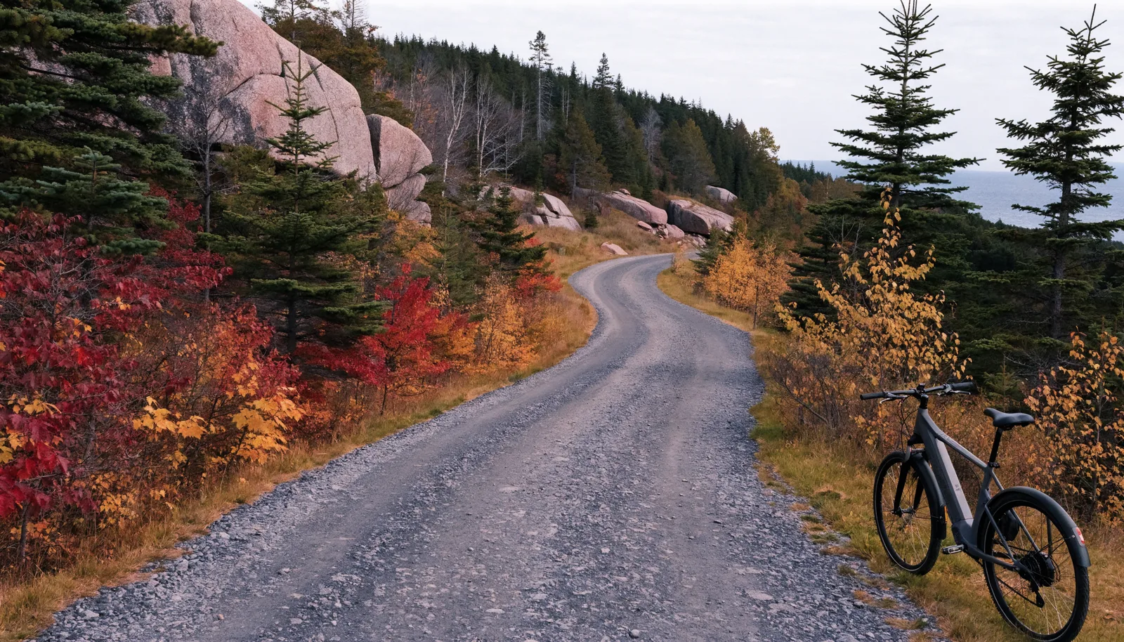 Class 1 electric bike on crushed-rock Acadia carriage road with fall foliage, granite hills, and a glimpse of the bay