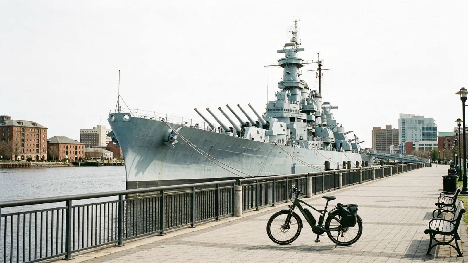 Electric bike on a paved waterfront promenade with railings beside a grey battleship museum ship moored at an urban river pier