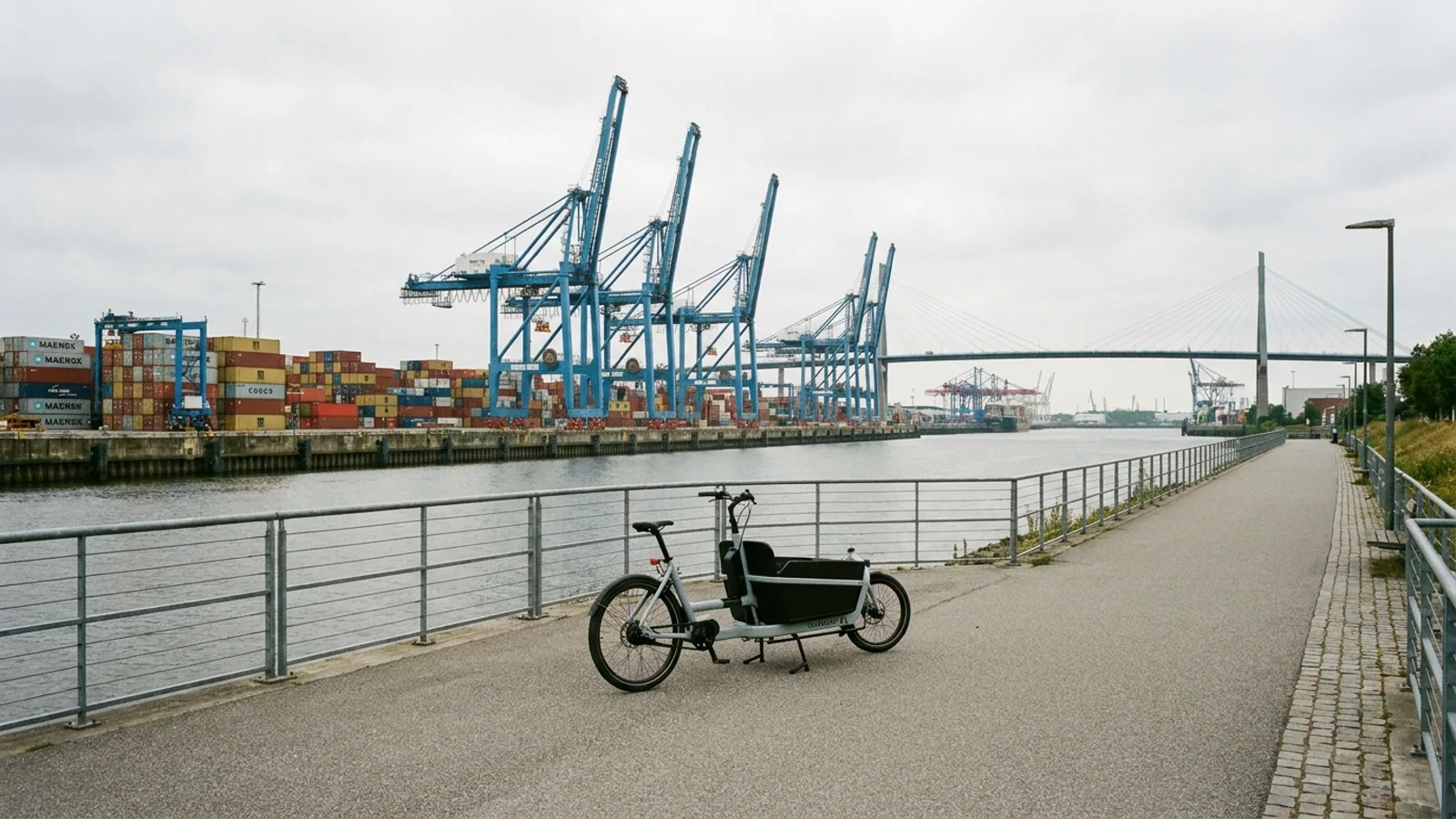 Electric bike on a paved waterfront path with railings looking across a channel to tall blue container gantry cranes and a long steel bridge
