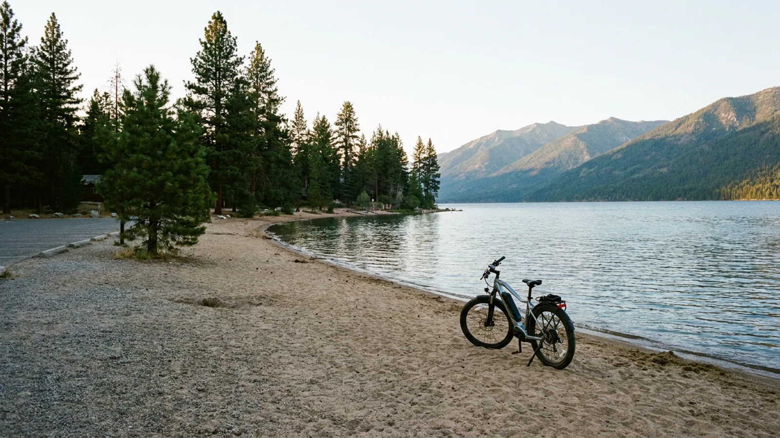 Electric bike at a quiet sandy state park beach on a large lake with mountains rising across the water and pines along the shore