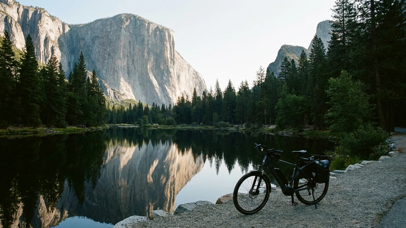 Electric bike on a gravel shore path beside a small dark mountain lake at the foot of a huge bare granite cliff reflected in still water