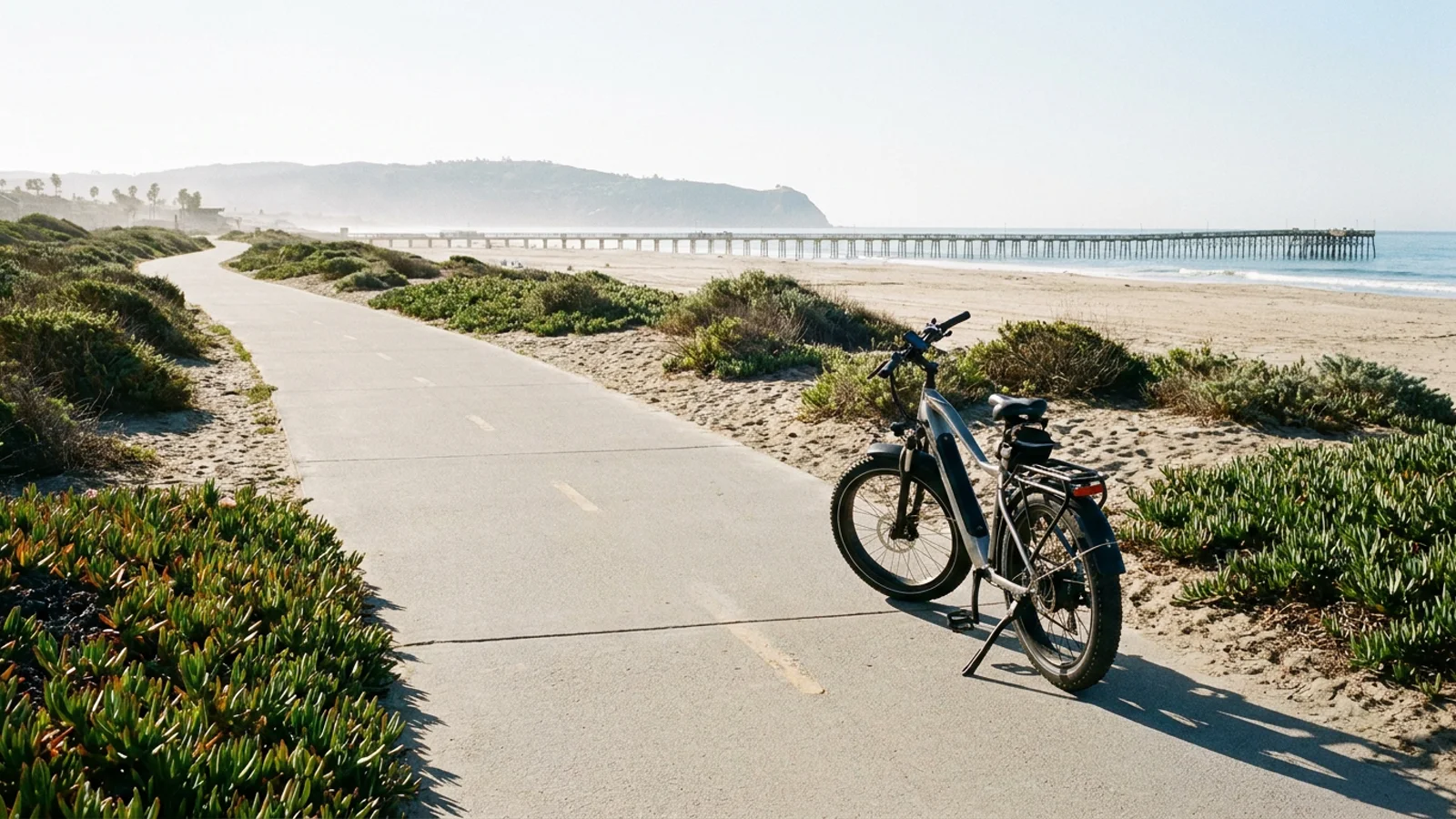 Electric bike on a wide paved beach bike path above the sand, with a pier and coastal headland in the distance