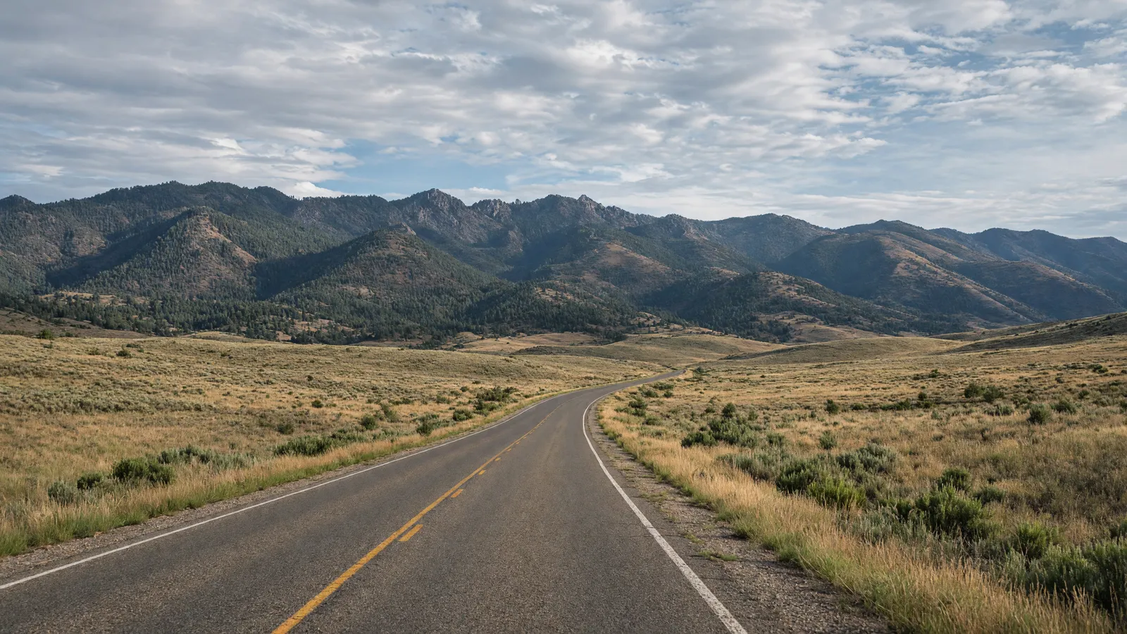 Empty Pecos River valley two-lane through high-country grassland toward forested mountains near Las Vegas, San Miguel County, New Mexico