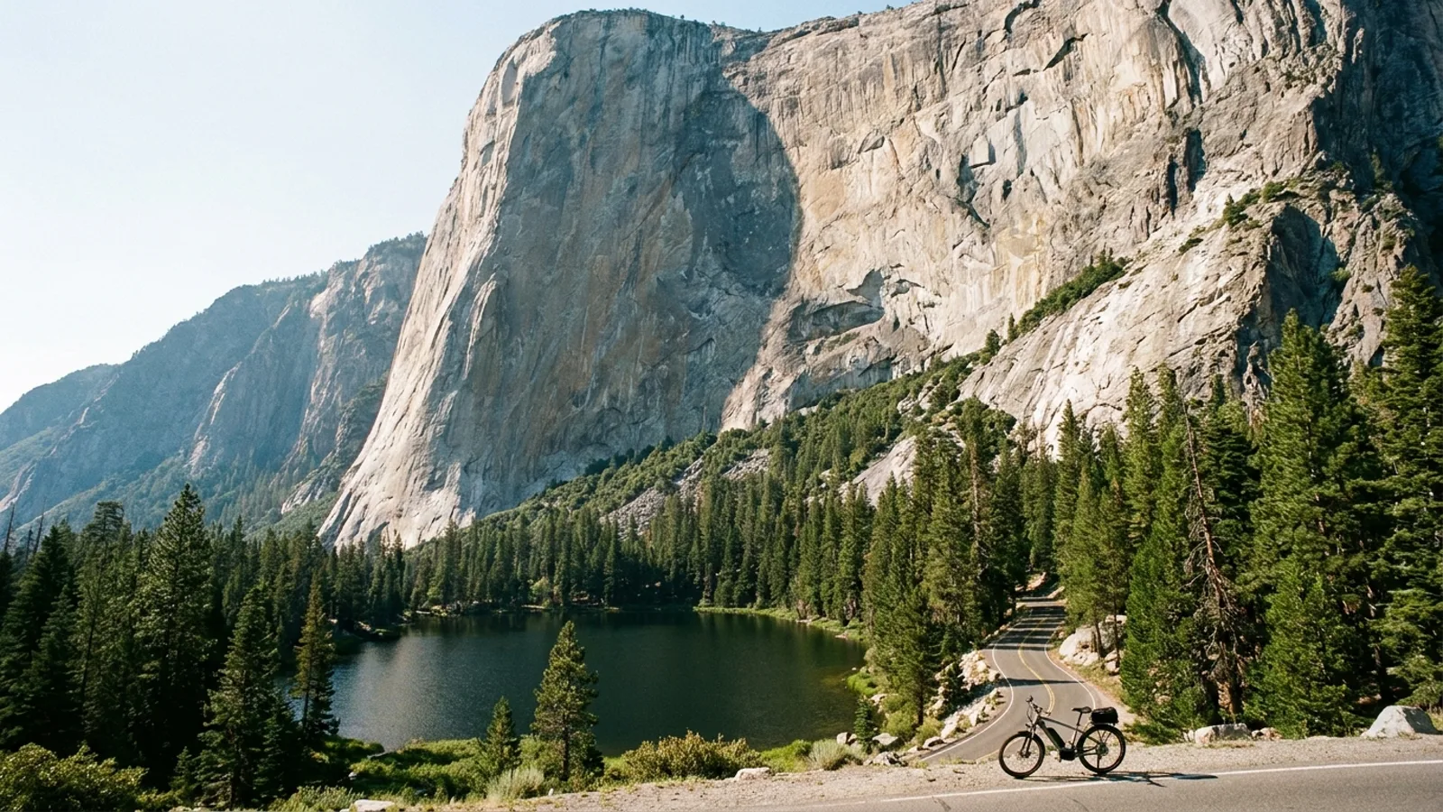 Electric bike at the roadside below a huge sheer granite cliff face rising above a small dark lake ringed with conifers, a narrow paved road switchbacking up at its base