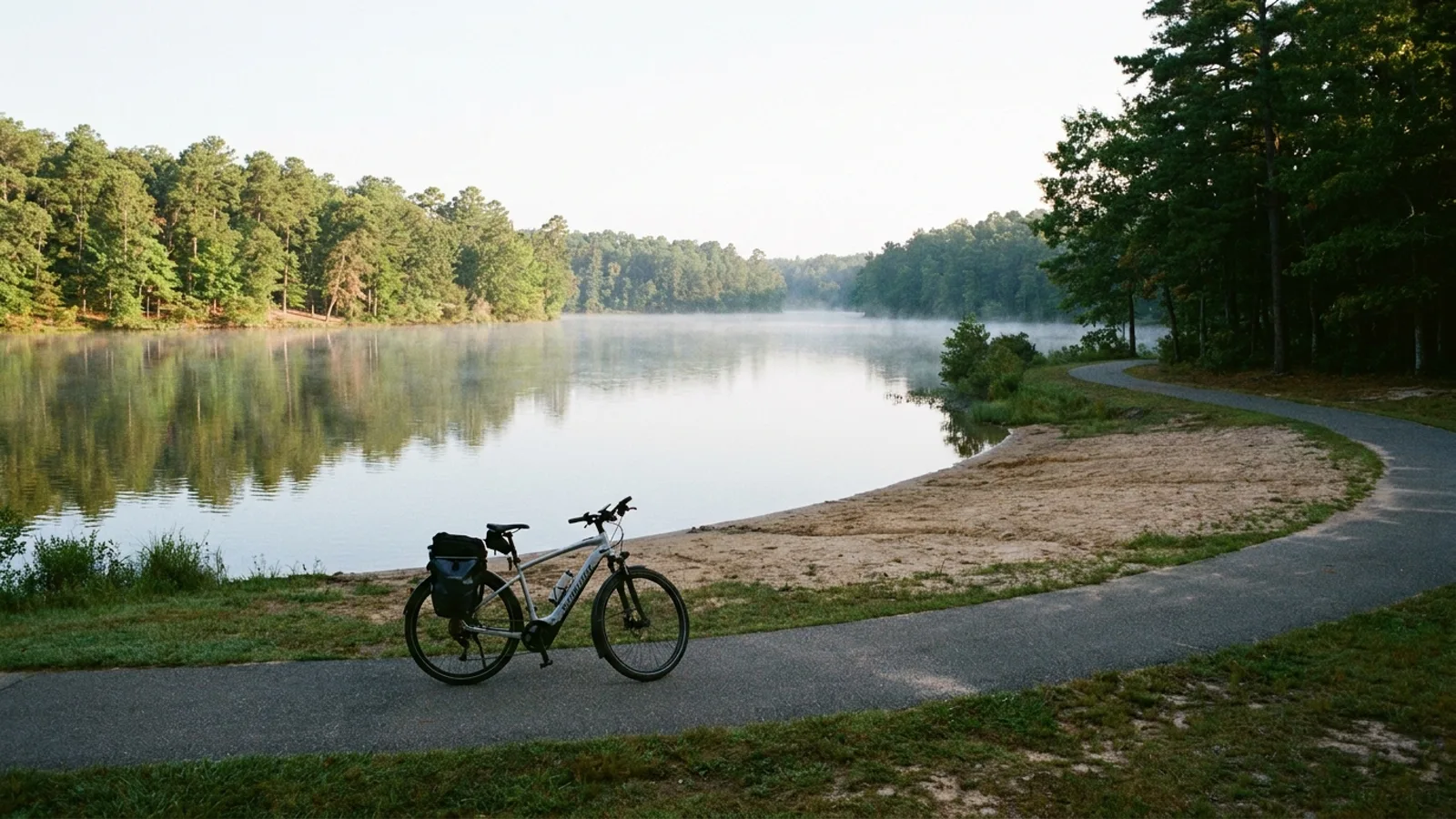 Electric bike on a paved loop path beside a calm lake ringed by pine and oak woods with a small sandy beach along one shore