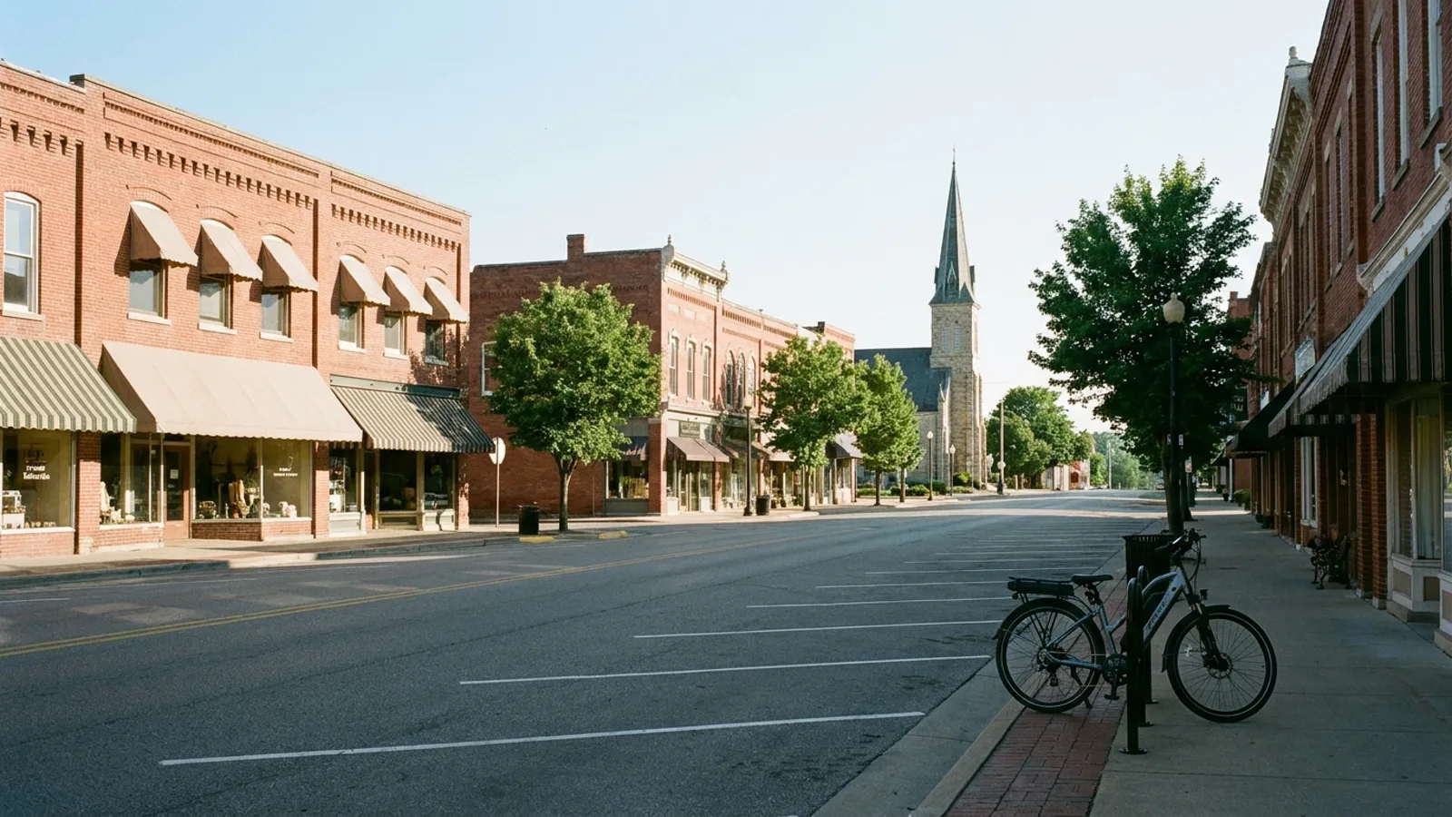 Electric bike at a rack on a wide historic small-town main street lined with brick storefronts and awnings with a tall church spire at the far end